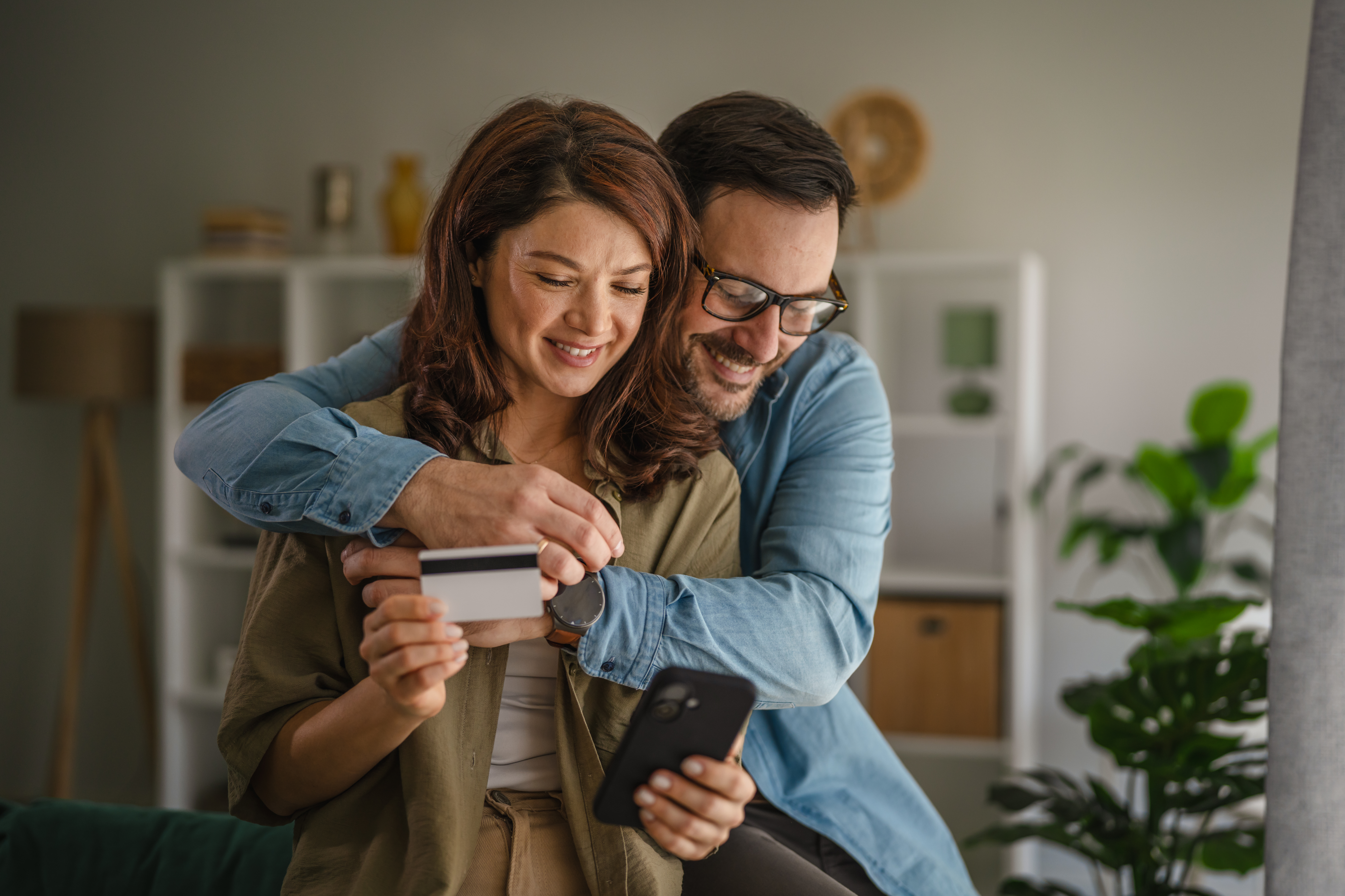 Smiling man embracing his partner while online shopping using a smart phone and holding a credit card, enjoying digital payment and secure e-commerce from the comfort of their home